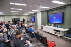 A large meeting room with the Astro logo showing on the monitors with dozens of people in chairs, waiting for the presentation to start.